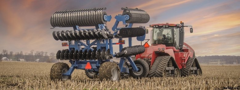 Folded blue Kinze Mach Till 302 pulled by a red Case IH tractor in a harvested corn field at sunset